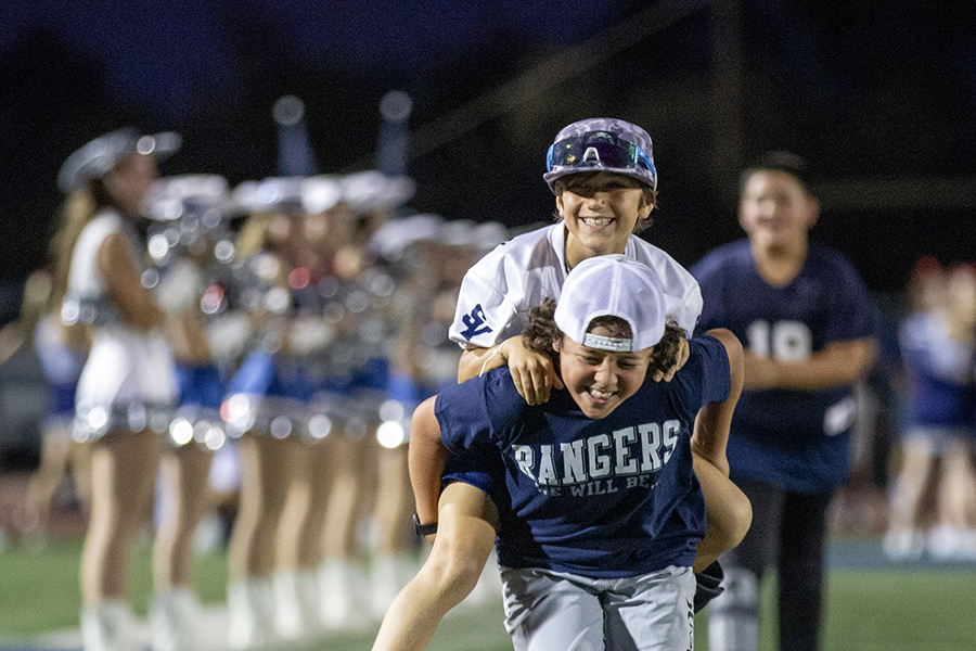 Youth league Ranger football players run down the field together. One player is carry his friend on his back because he is in a boot and cannot run.